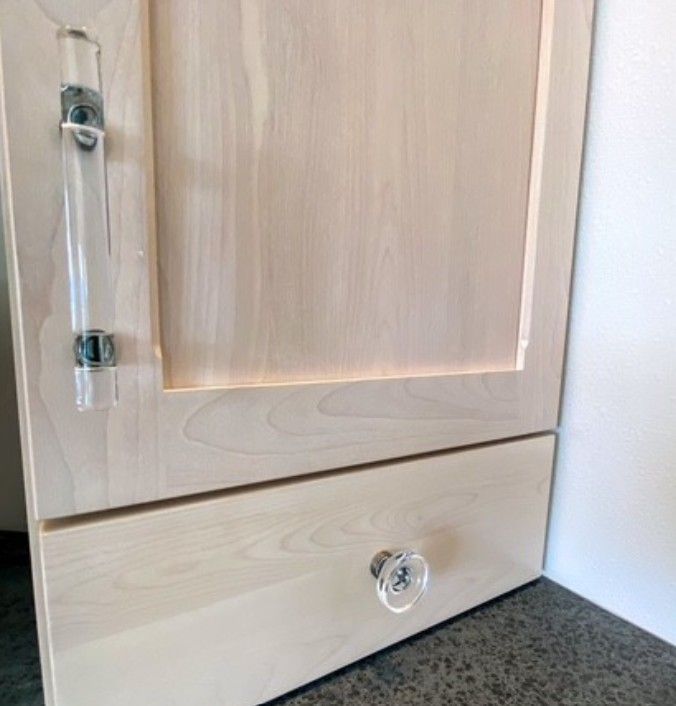 Light wood cabinet with a clear acrylic handle and a small bottom drawer, beside a white wall and gray carpet.