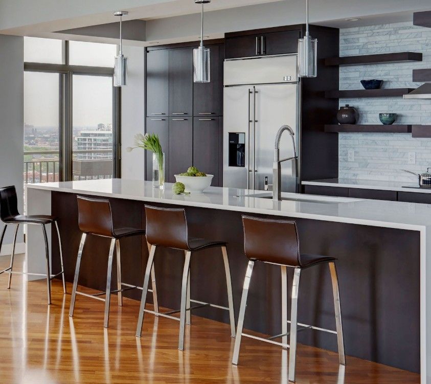 A modern kitchen with a long white-topped island, dark cabinetry, stainless steel appliances, and four brown bar stools.