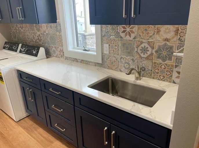 Kitchen with navy cabinets, patterned backsplash, white countertop, and stainless sink below a window