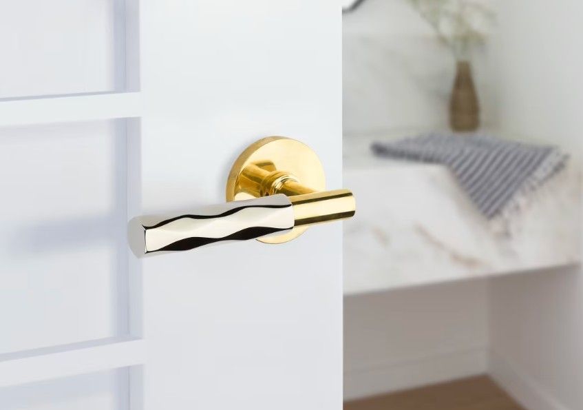 A gold-toned, modern door handle with a textured, wavy silver finish, attached to a white interior door.