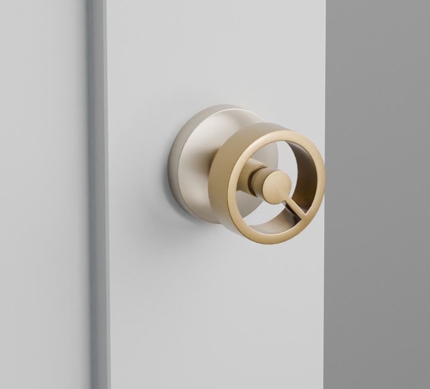 A circular, gold-toned door knob with a modern ring design, mounted on a light gray door panel against a gray background.
