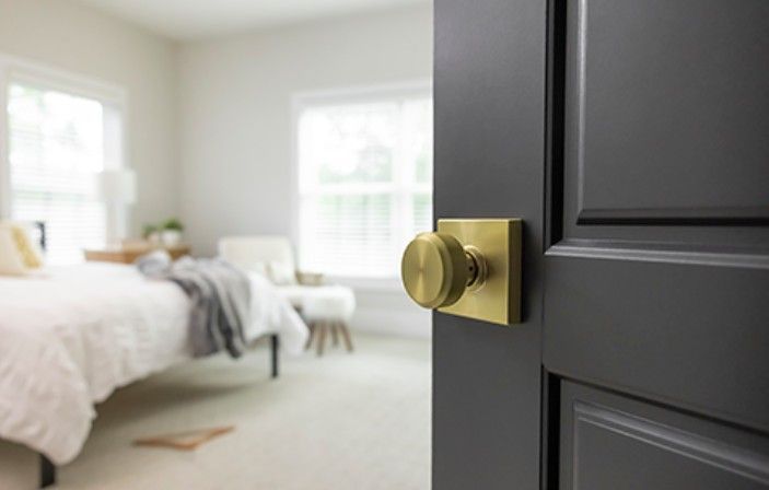 A brass door knob on a dark grey door, partially opened to reveal a blurred, bright, and airy bedroom in the background.