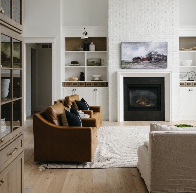A bright living room with tan leather chairs, a white fireplace with a TV, built-in shelving, and light wood floors.