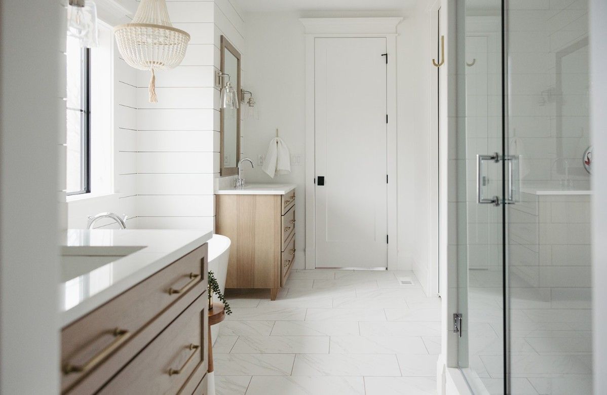 A bright, modern bathroom with light wood cabinets, white walls, white tile floors, and a glass-enclosed shower.