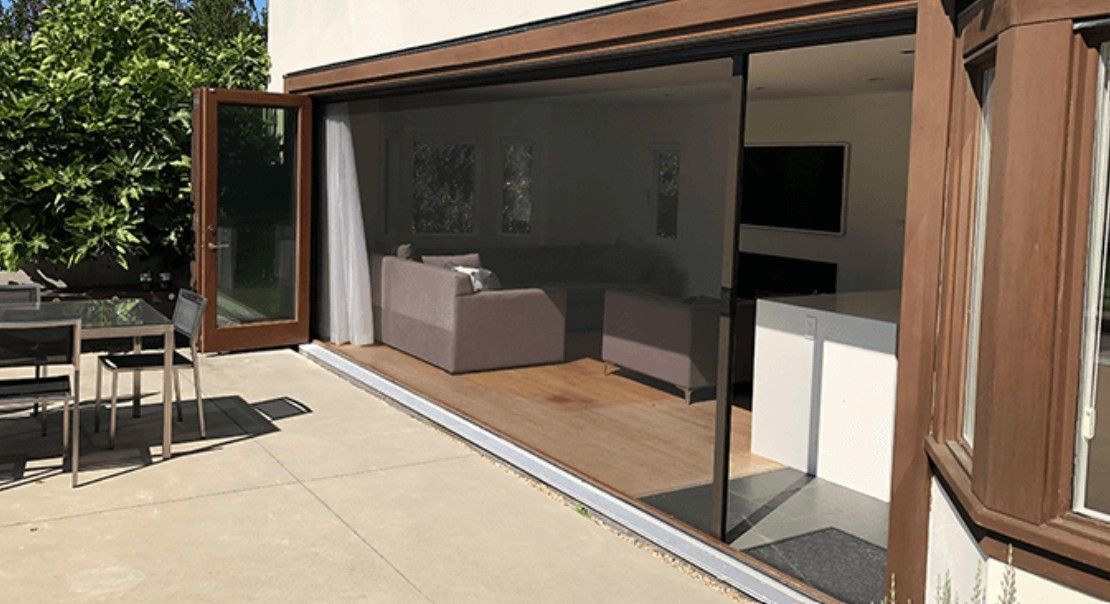 Modern patio with open sliding glass doors to a living room, featuring outdoor seating and a wooden deck