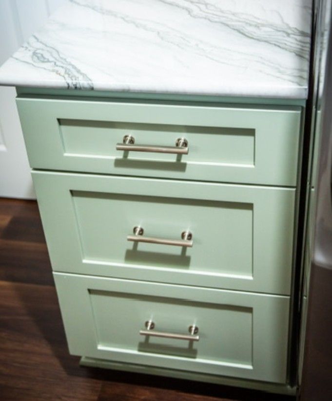 Mint green kitchen cabinet with three drawers and silver handles under a white marble countertop