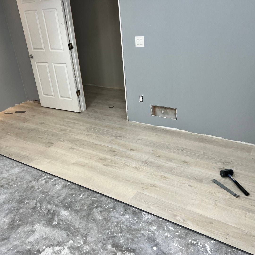 Laying light wood-look flooring in a room with a partially finished concrete floor, doorway, and gray walls.