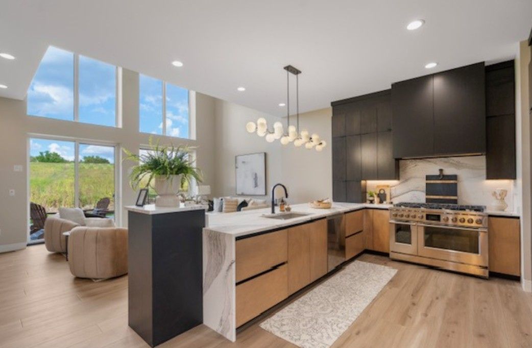 Modern open-concept kitchen with wood cabinetry, white island, pendant lights, and large windows overlooking greenery