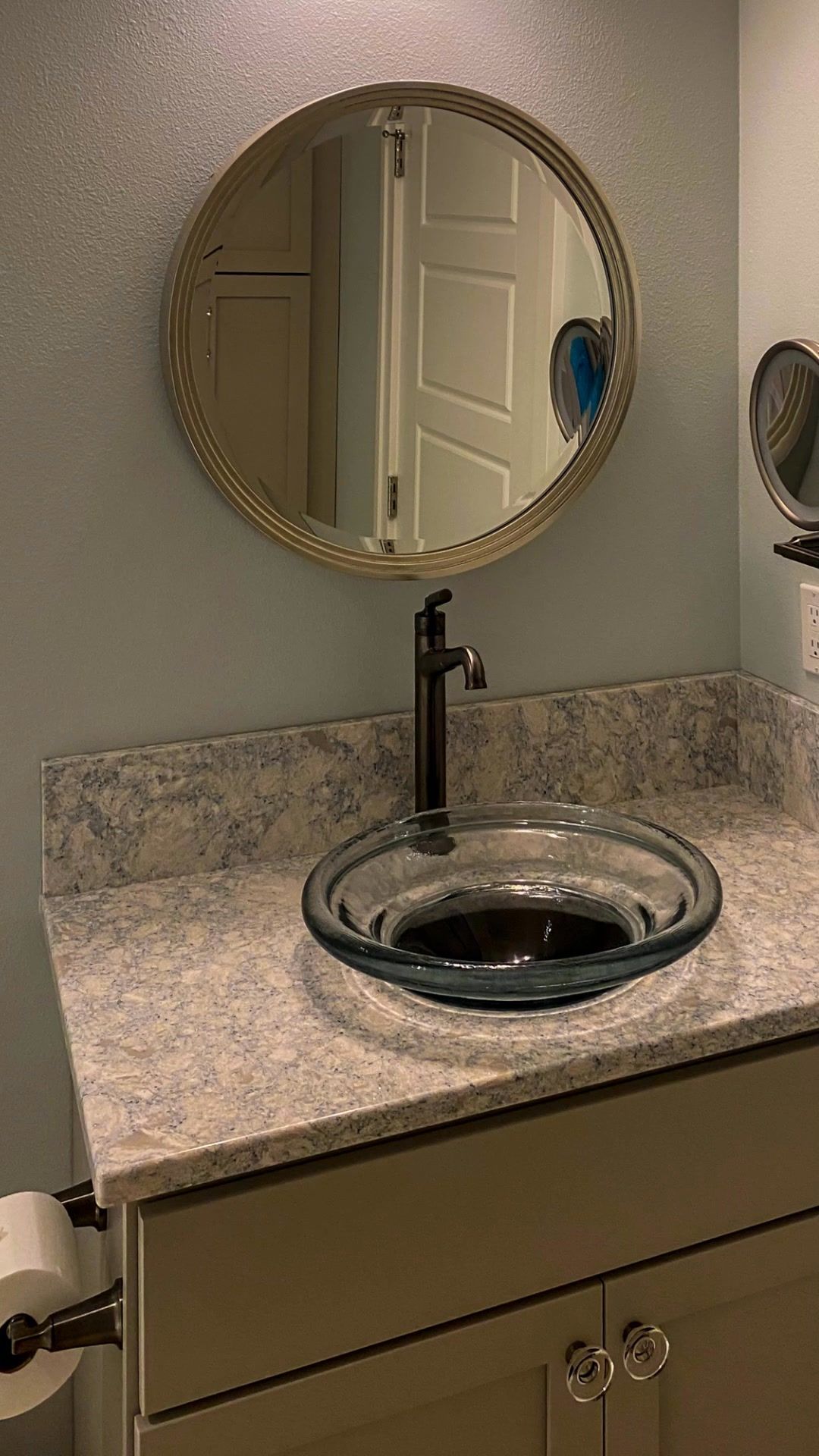 Bathroom with round mirror, glass sink, dark faucet, and light-colored countertop and cabinetry.