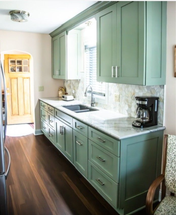 Narrow kitchen with sage-green cabinets, white countertops, and dark wood floor, with daylight from a window