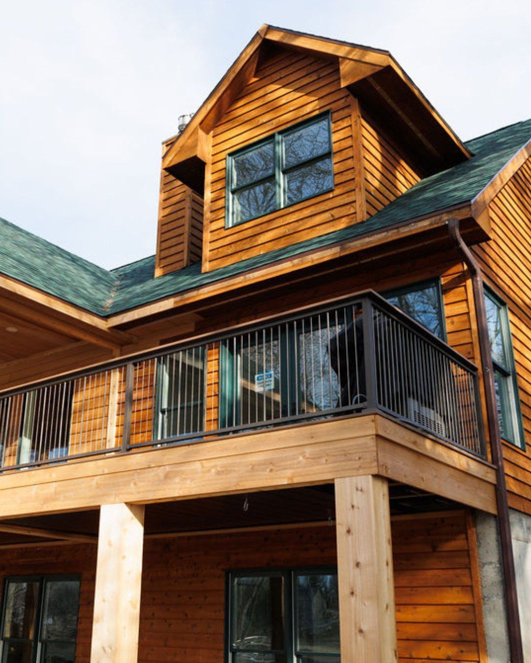 Wooden house with a deck and dark railings, green roof, and clear blue sky.