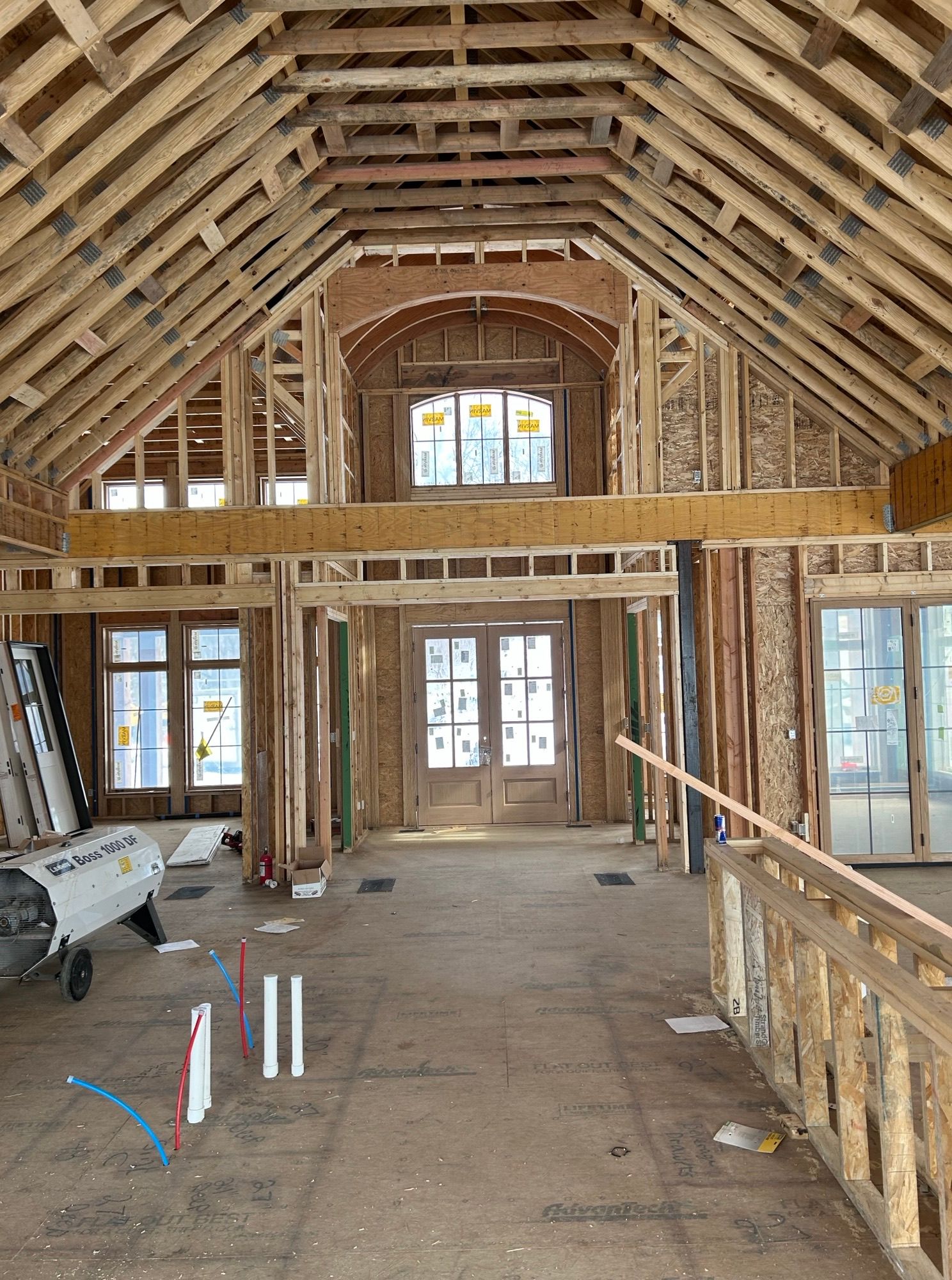 Interior of a house under construction. Wooden framework, unfinished floors, arched window, double doors, and heater visible.