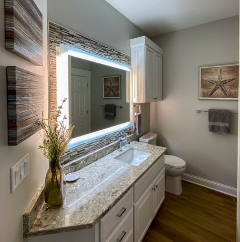 Bright bathroom vanity with lit mirror, marble countertop, white cabinets, and wood floor