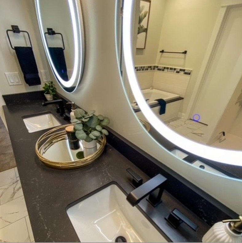 Modern bathroom vanity with round LED mirrors, black countertop, white sinks, and gold tray decor