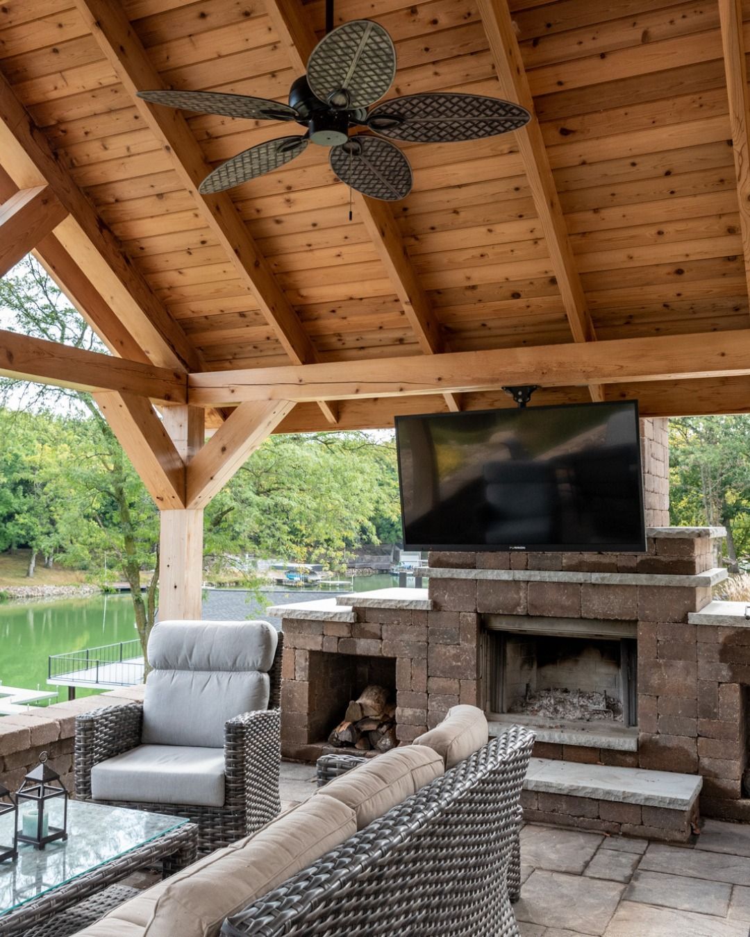 Outdoor seating area with fireplace, TV, and ceiling fan under a wooden pergola, overlooking a lake.