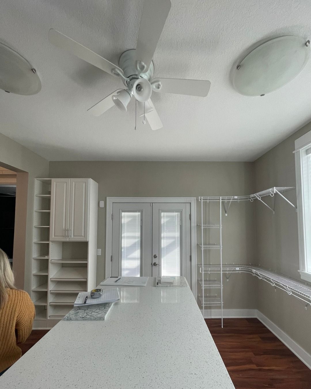Kitchen interior: white island, cabinets, ceiling fan, and double doors to outside, with someone standing near the island.