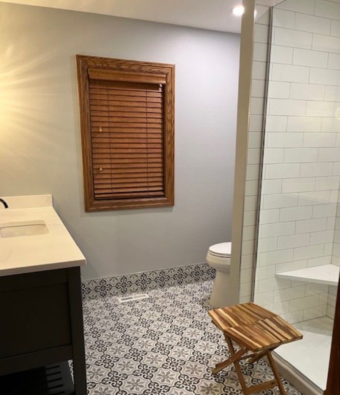 Small bathroom with patterned tile floor, wooden stool, shower, toilet, vanity, and window blinds