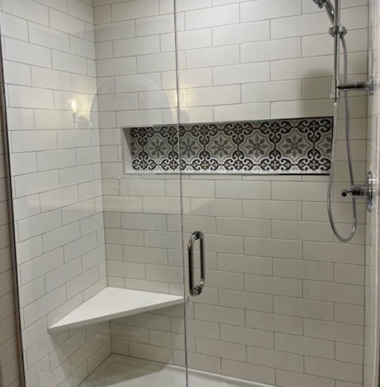 White tiled shower stall with built-in corner bench, grab bar, handheld shower, and decorative tile band