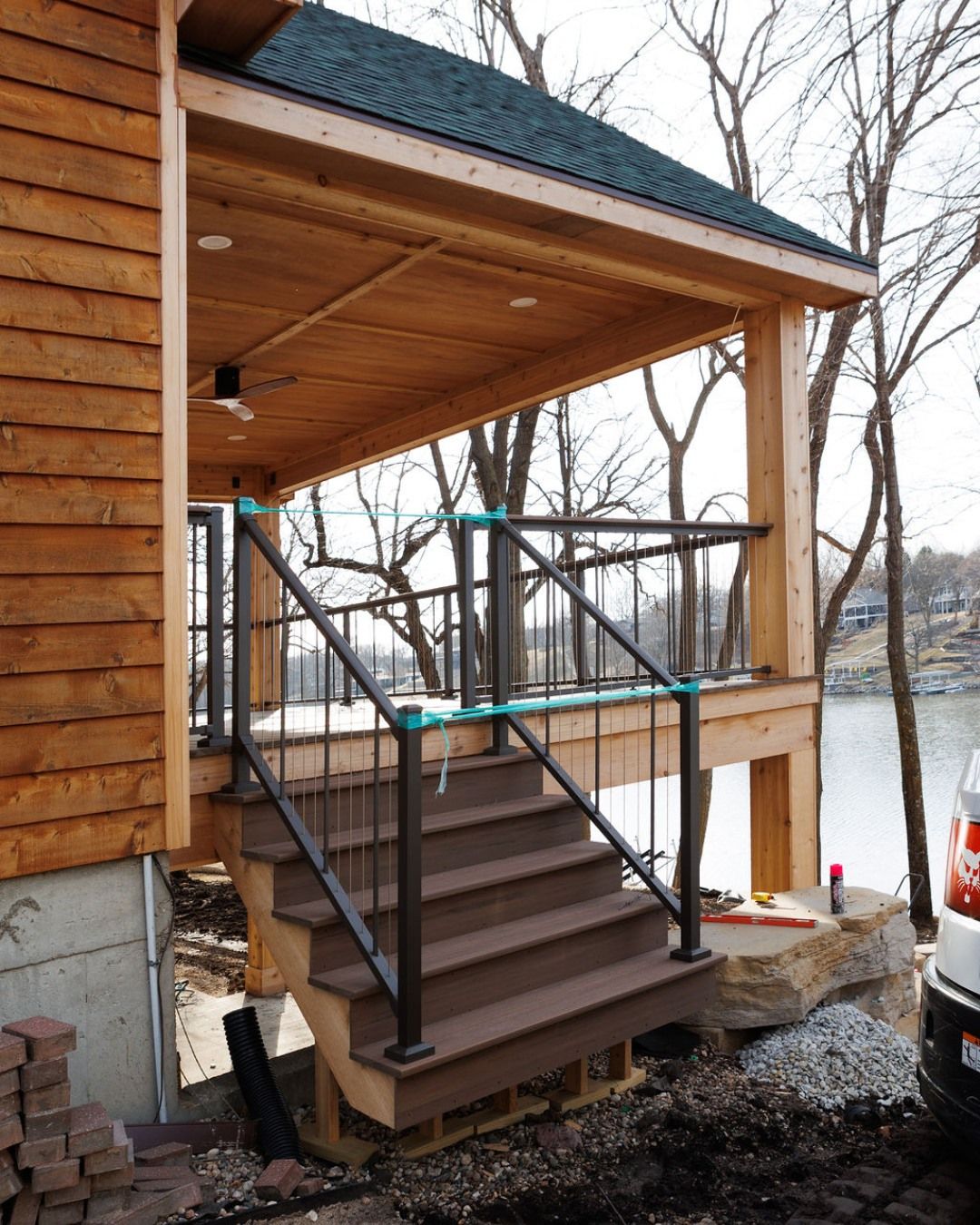 A wooden porch with stairs leading down; dark metal railings, overlooking a lake.