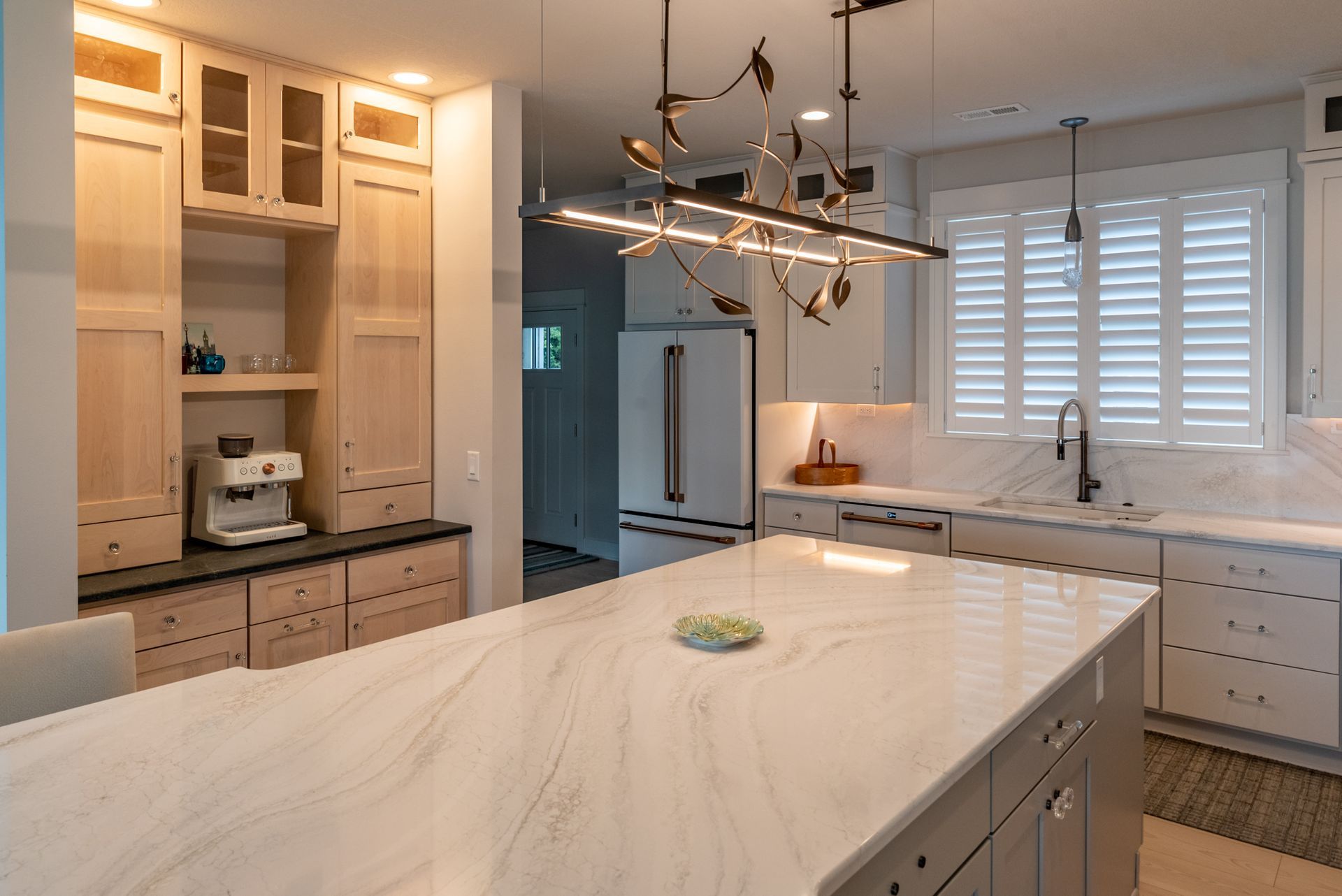 Modern kitchen with light wood cabinets, marble island, stainless steel appliances, and large window with blinds.