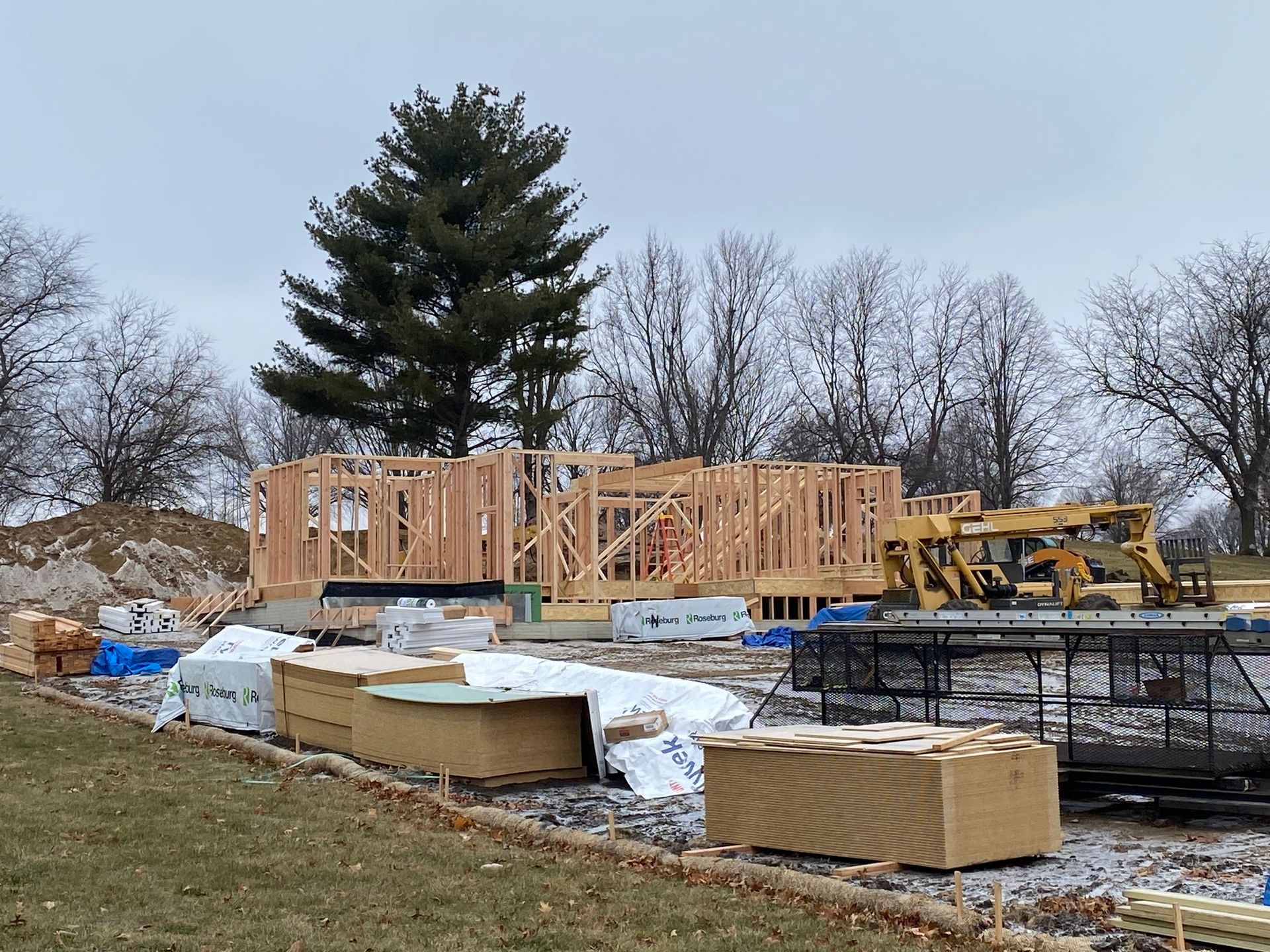 Framed wood structure of a house under construction; construction materials and machinery are visible on a cloudy day.
