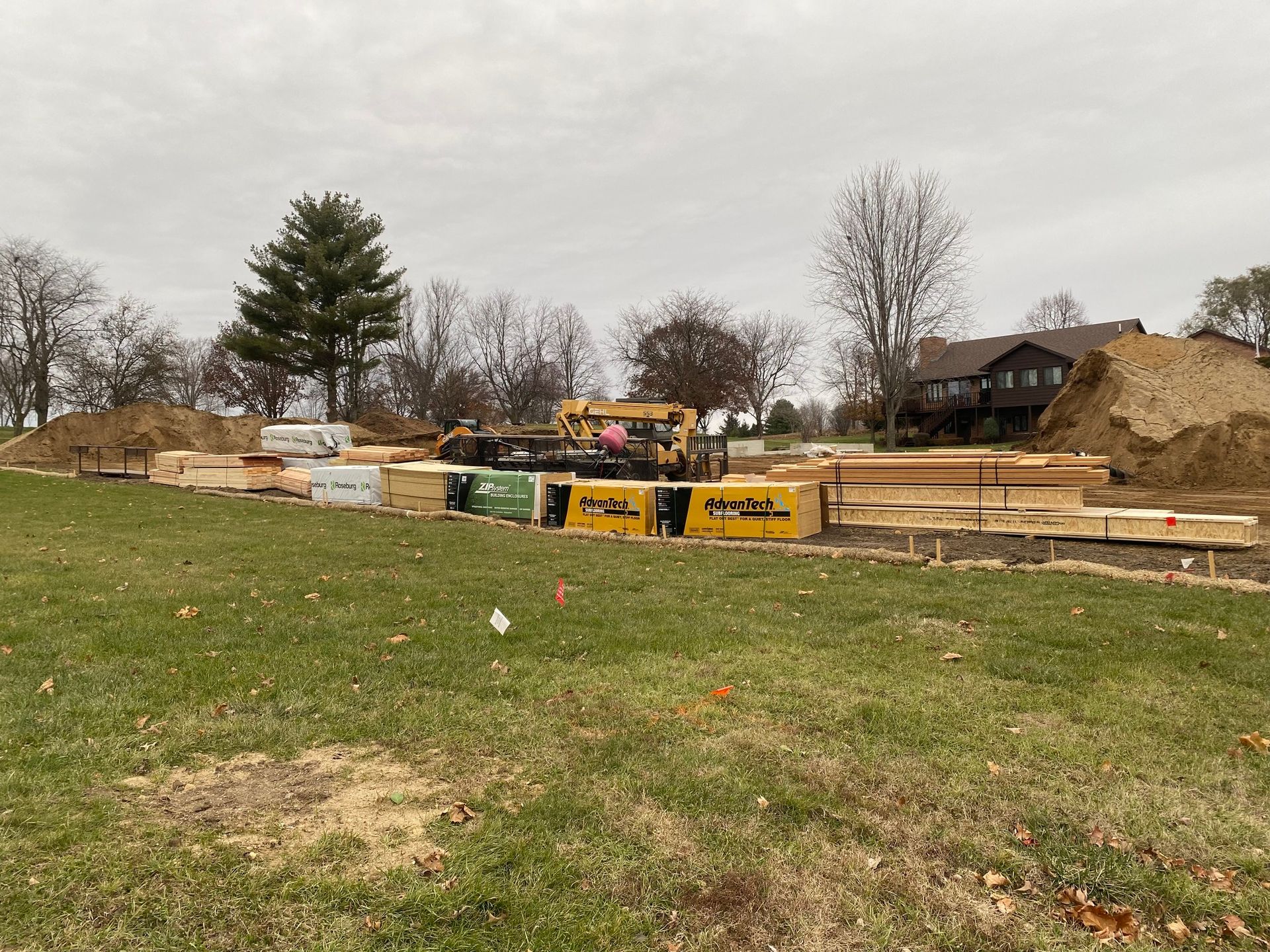 Construction site with lumber and heavy machinery, a house in the background. Overcast sky, grass in the foreground.