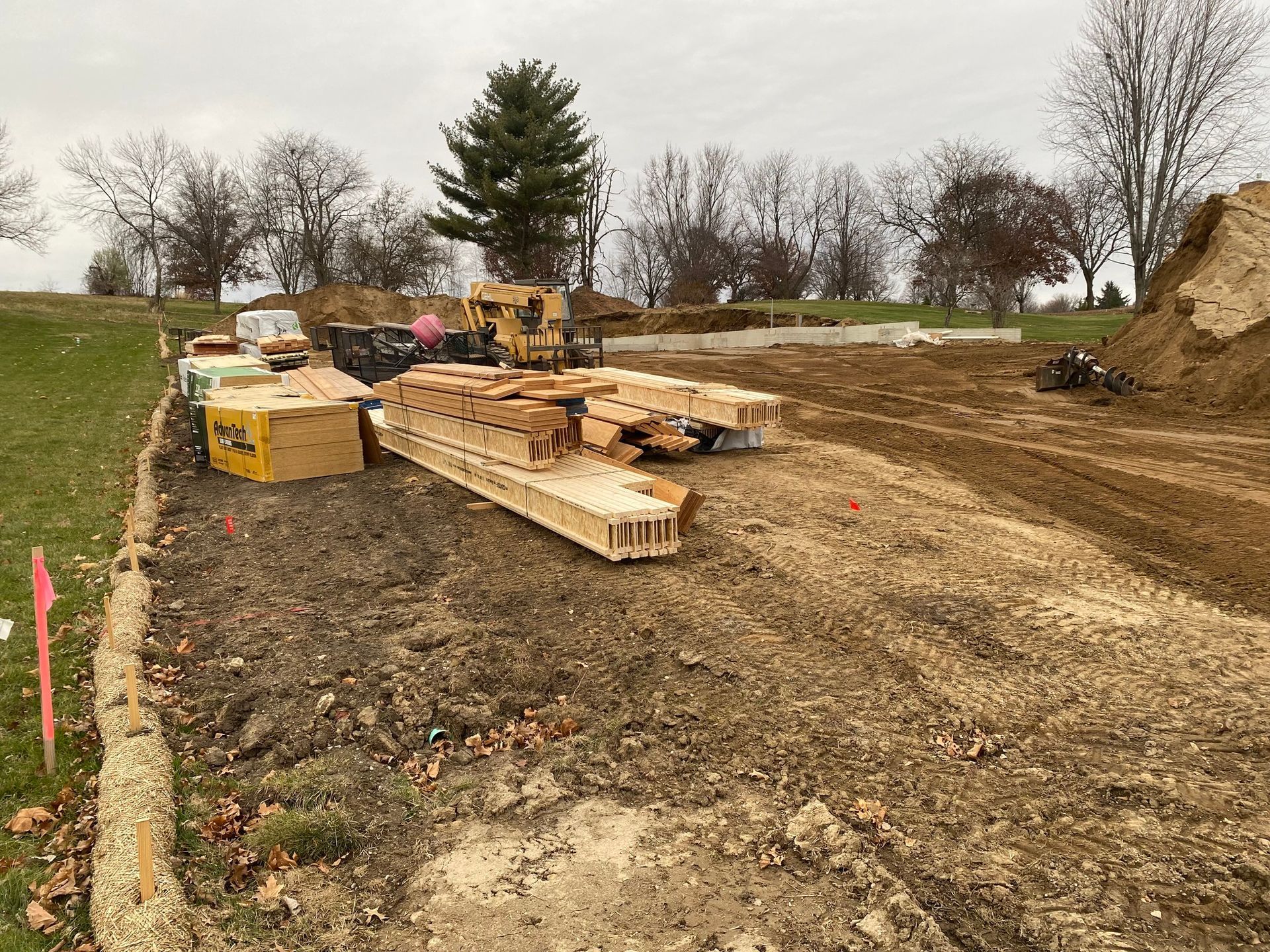 Stacks of wooden construction materials sit on a dirt lot at a building site, with an excavator in the background.