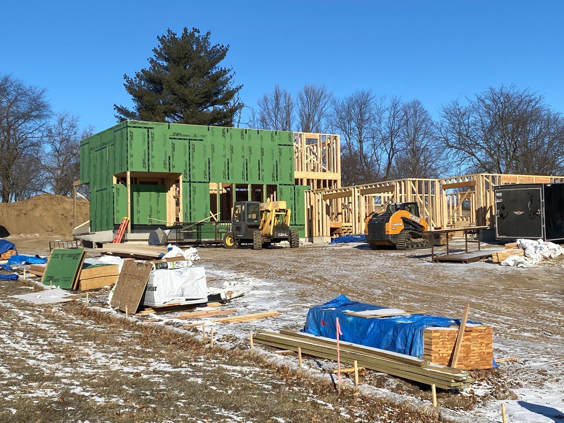 Construction site with a partially framed house, green sheathing, yellow heavy machinery, and scattered building materials.