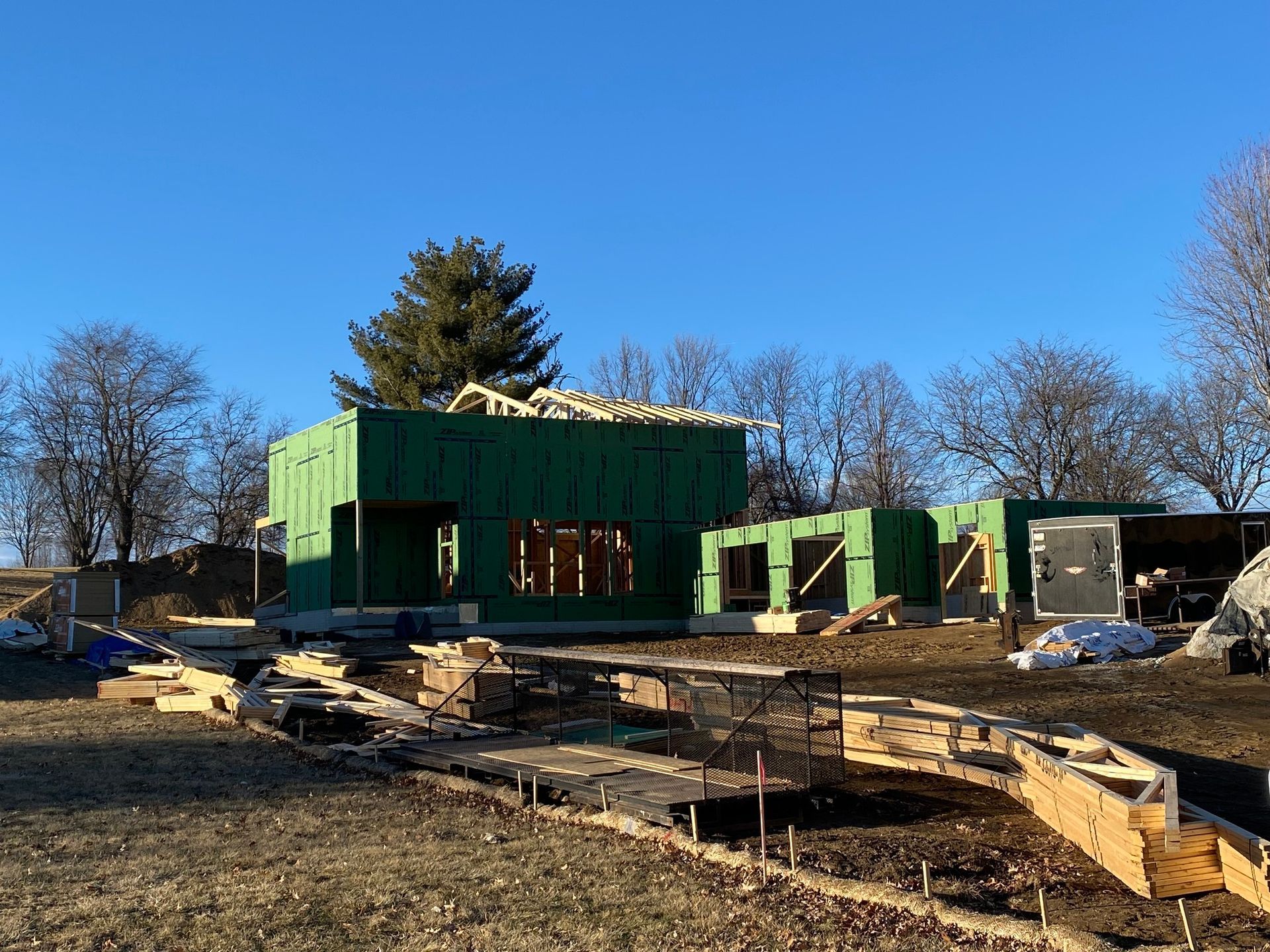 A house under construction with green exterior wall panels, wooden framing, and exposed ground on a sunny day.
