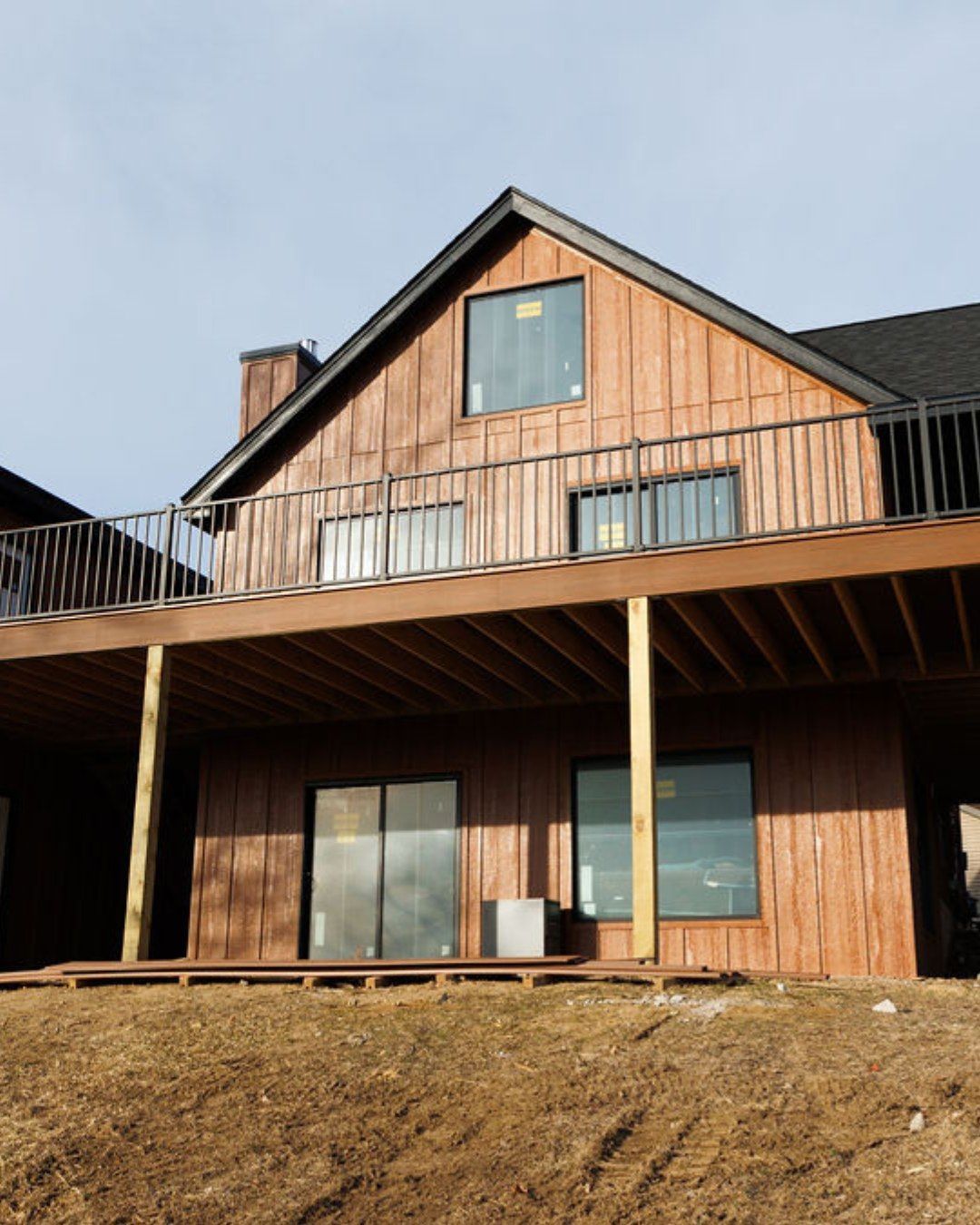 Brown house with deck, black railings, and windows on a hillside, against a cloudy sky.