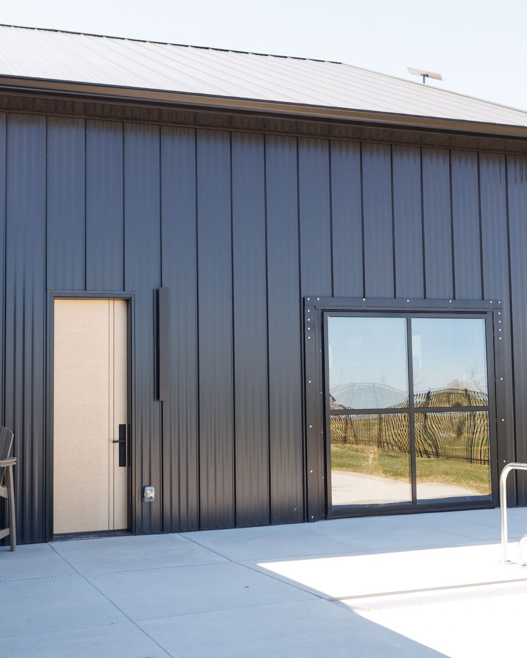 Black building with a door and a sliding glass door, reflecting a vineyard. Concrete patio in the foreground.
