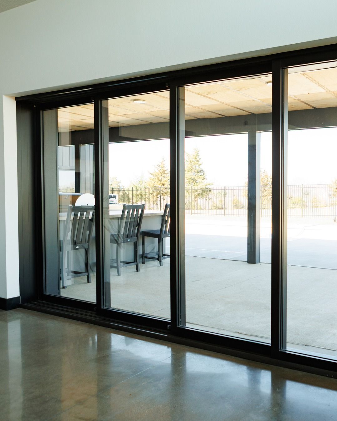 Black-framed sliding glass doors open to a patio with concrete flooring, chairs, and an outdoor view.