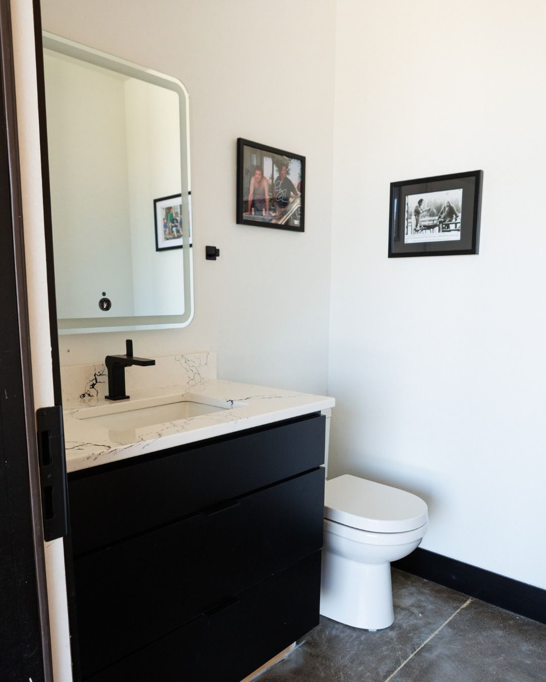 Modern bathroom with black vanity, white countertop, and white toilet. Includes mirror and framed photos on white walls.