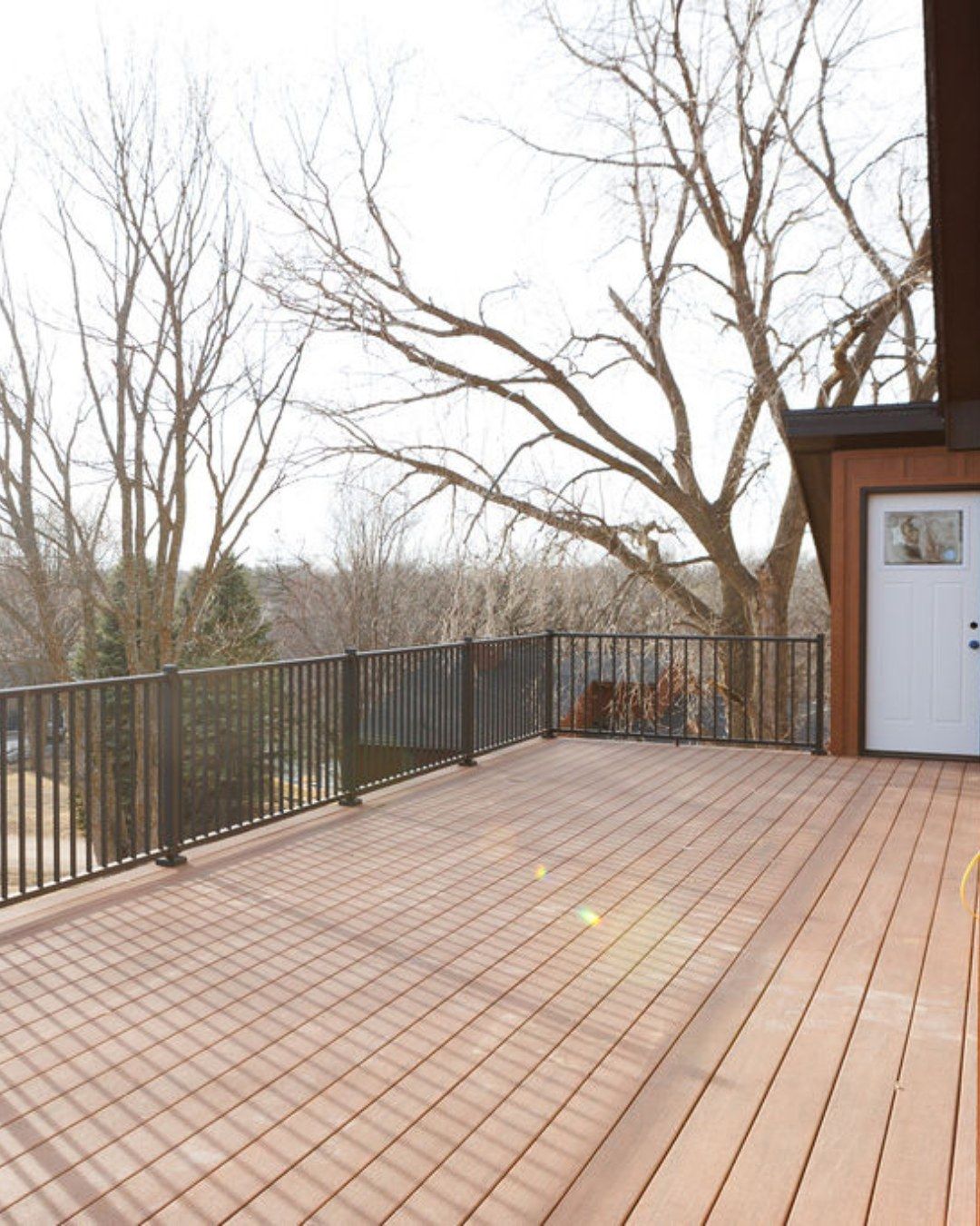 Wooden deck with black railings and a white door, surrounded by trees.