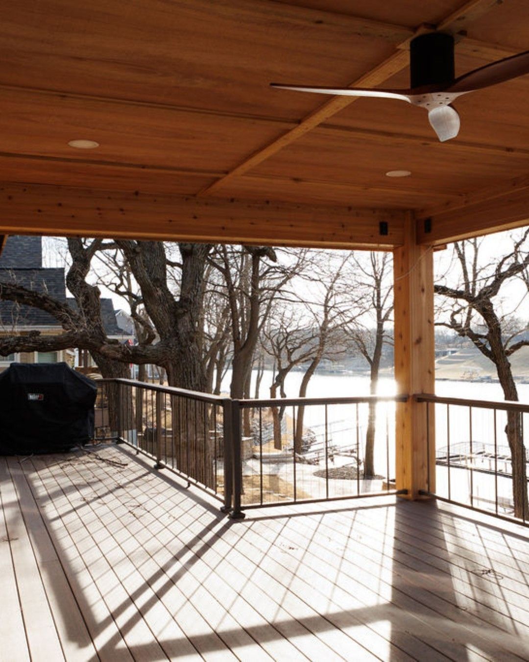 Covered wooden deck overlooking a lake with trees and a black grill.