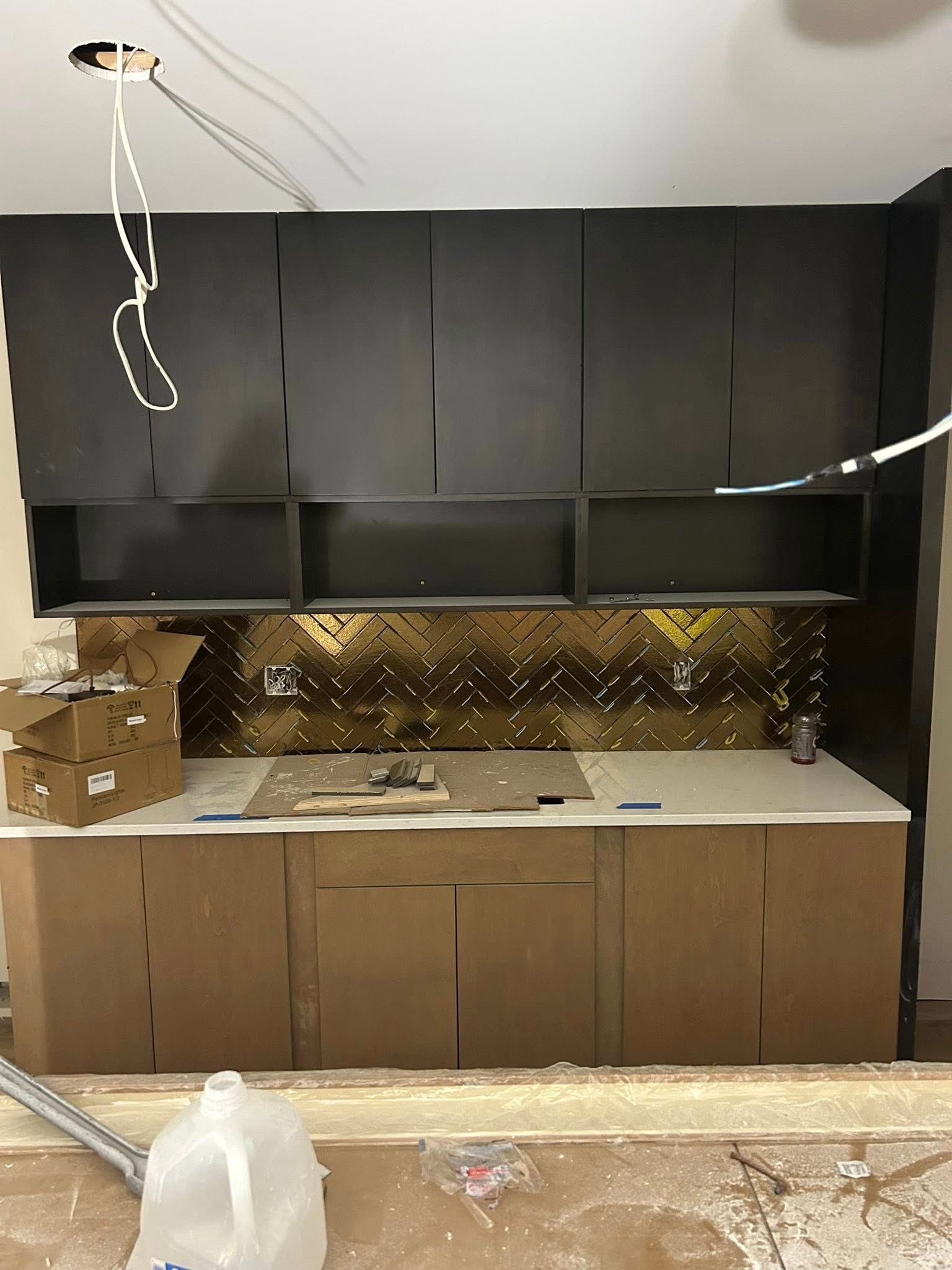 Kitchen under construction with dark upper cabinets, gold backsplash, and light lower cabinets.