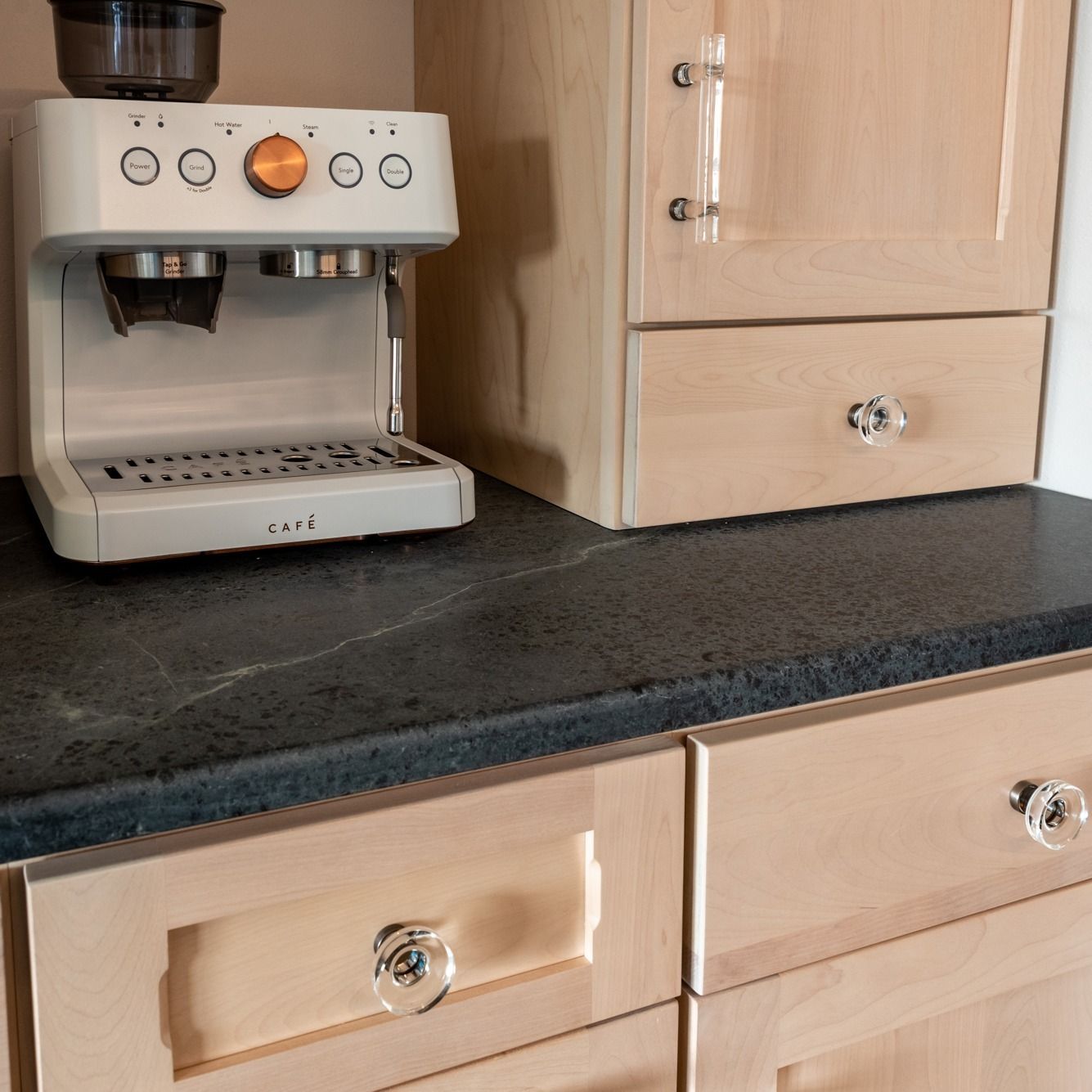 Espresso machine on a dark countertop next to light-colored cabinets with clear knobs.