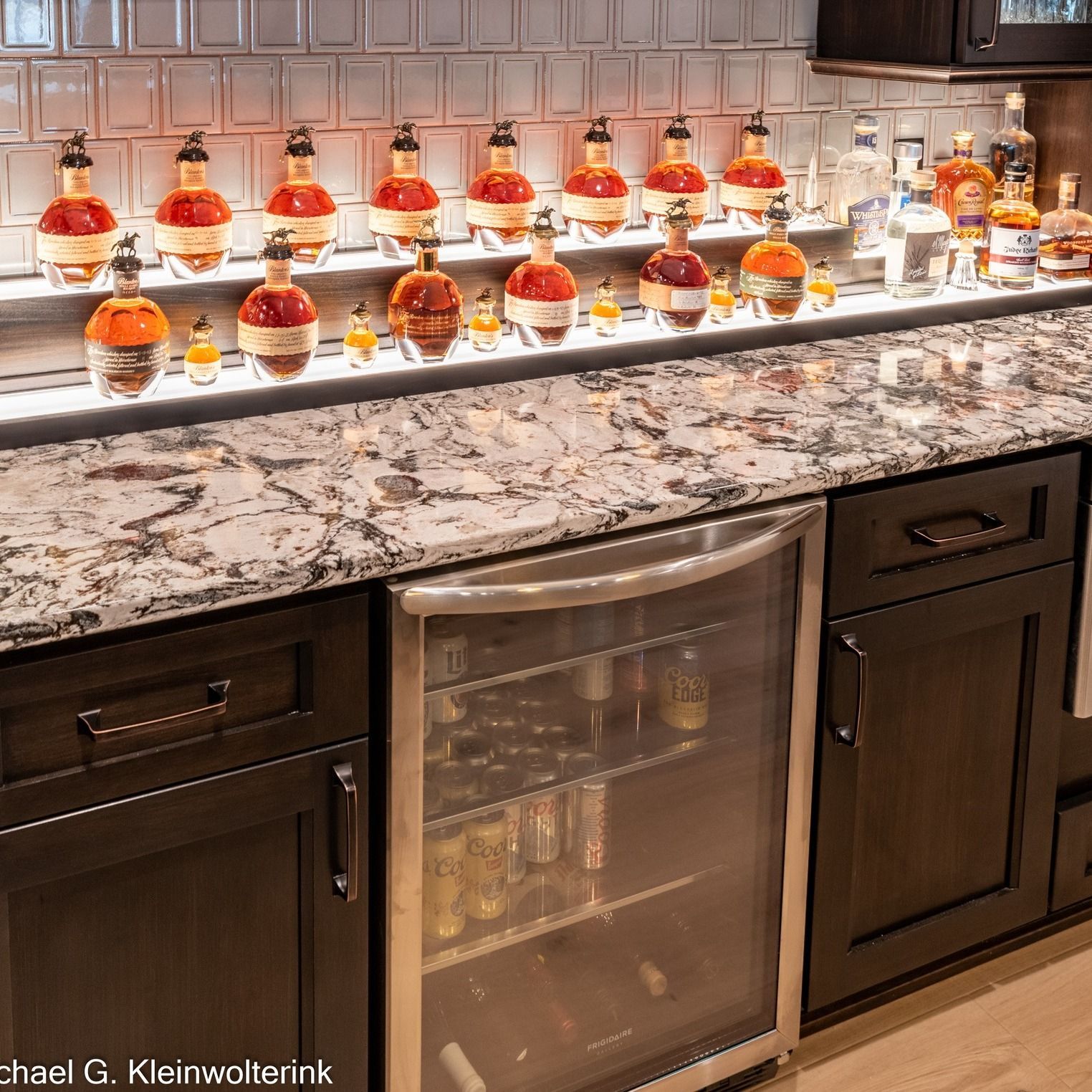 Bar with bottles displayed on shelves, granite countertop, and small refrigerator.