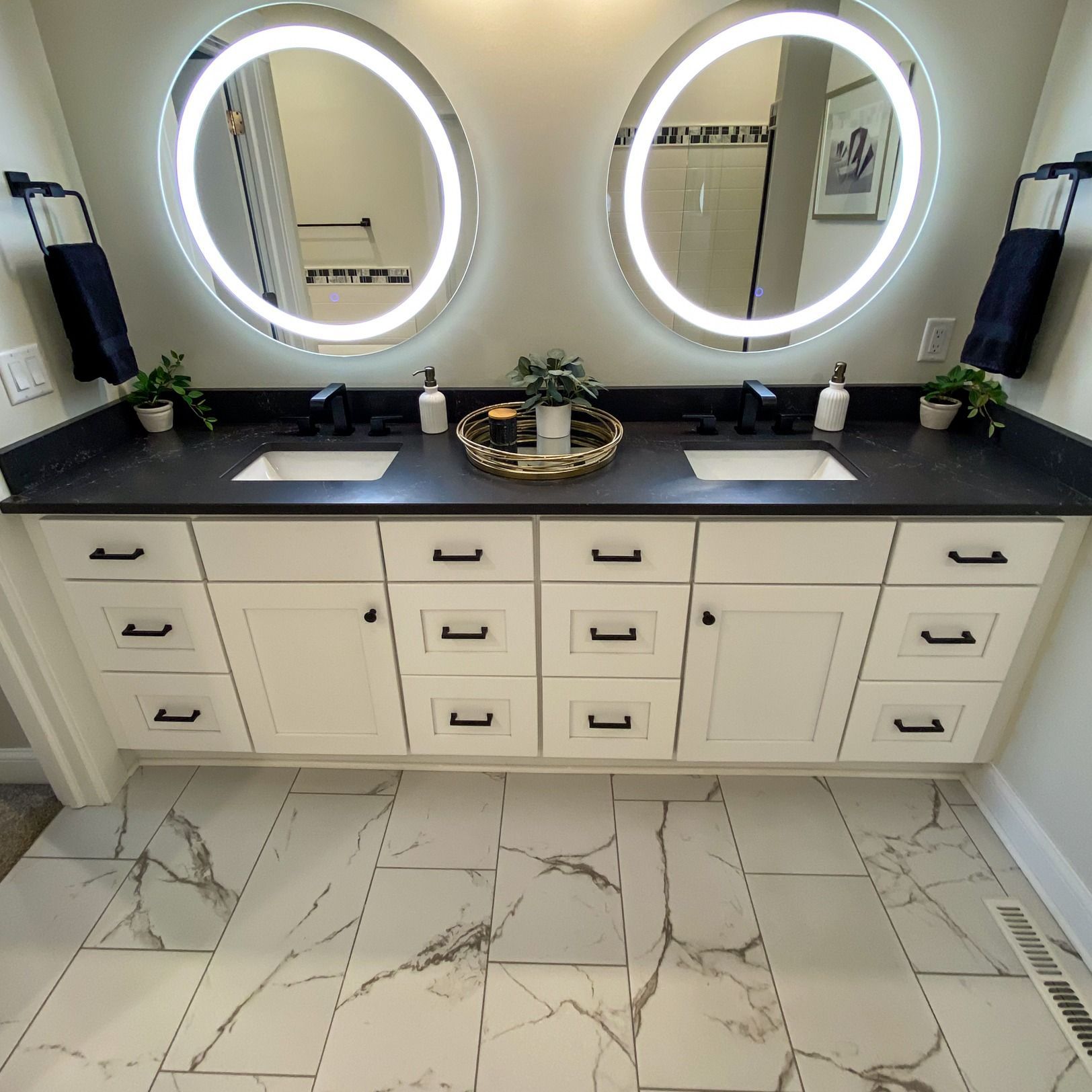 Bathroom with double sinks, white vanity, black countertop, lit oval mirrors, and marble tile floor.