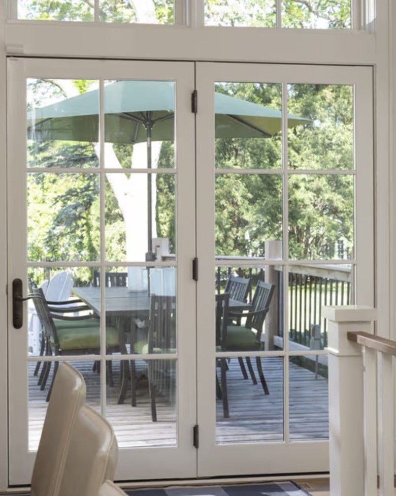 White double French doors look out onto a deck featuring a patio dining set with a green umbrella and lush trees.