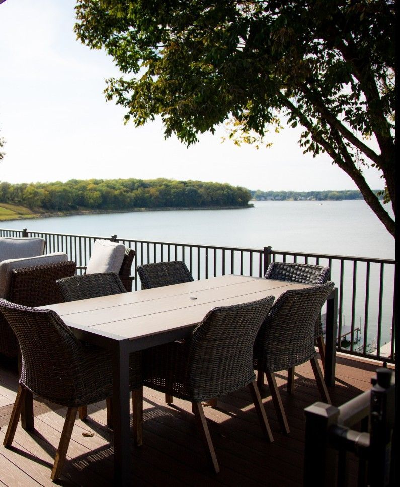 Wicker patio dining set on a deck overlooking a lake with a tree overhead.