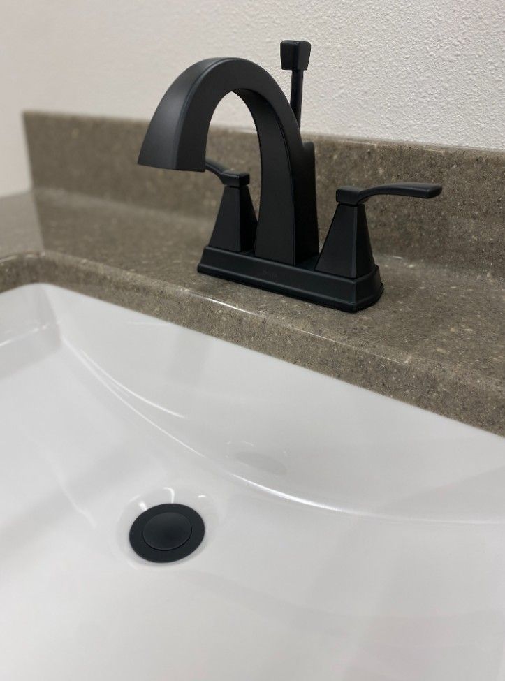 A matte black bathroom faucet with two handles and a matching drain stopper, mounted on a grey stone vanity countertop.