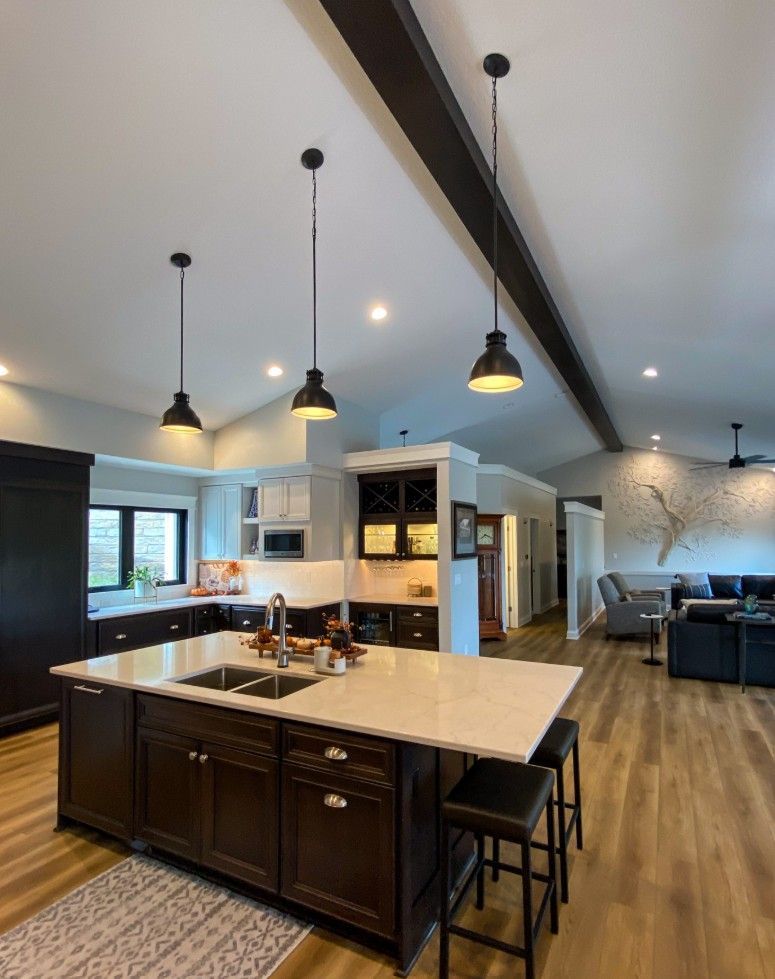 A modern kitchen with a dark brown island, white countertops, hanging pendant lights, and wood floors in an open floor plan.