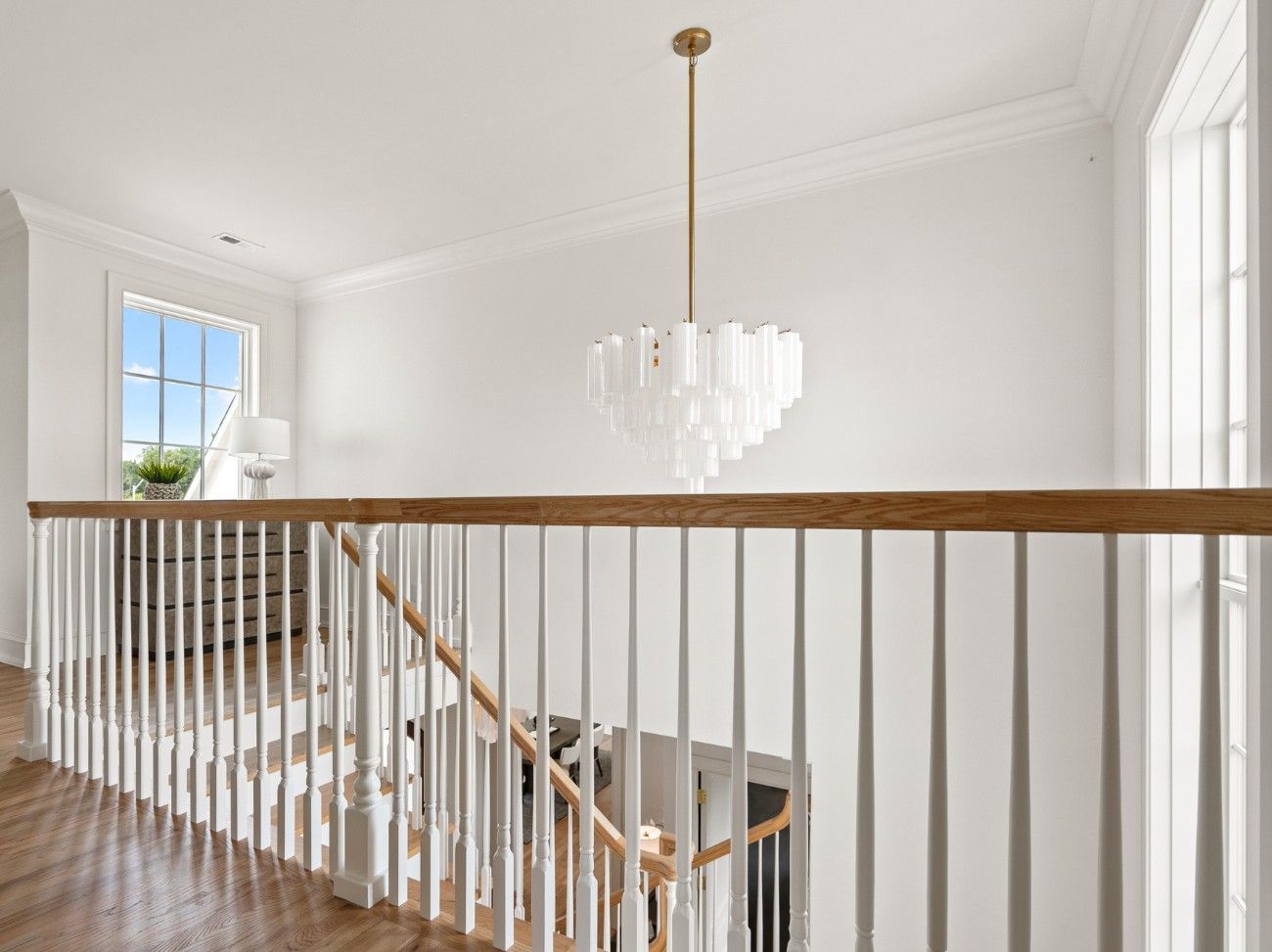 A bright indoor landing features white railings, a wood handrail, and a modern tiered chandelier hanging above the stairs.