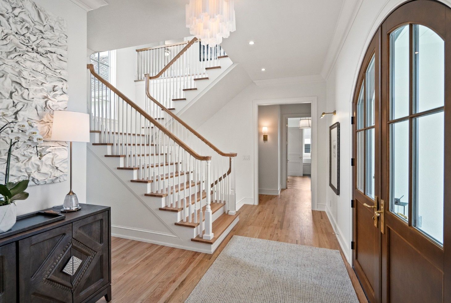 A bright foyer with white walls, a wooden staircase, a dark console table, a patterned rug, and double wooden front doors.