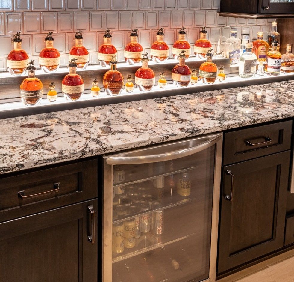 A home bar with granite countertops, dark wood cabinets, a wine fridge, and two lit shelves displaying many liquor bottles.