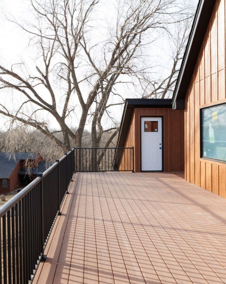A wide wooden deck with a black metal railing, featuring a white door on a house with vertical brown siding near trees.