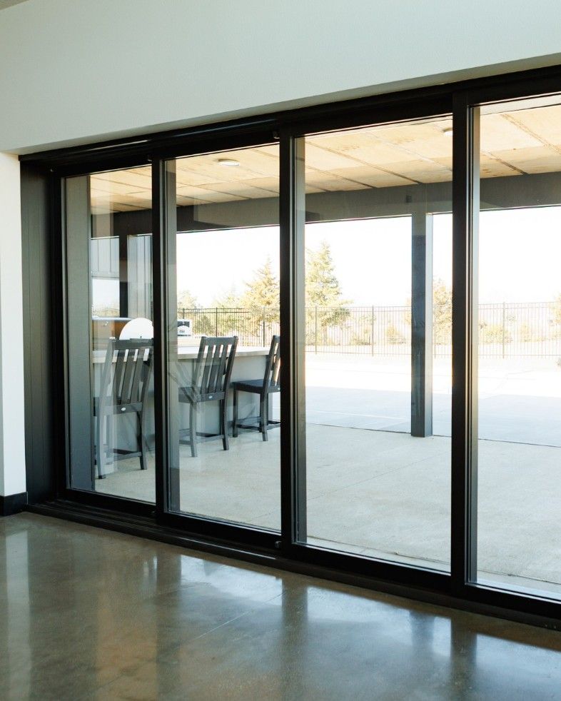Black-framed sliding glass doors open onto a concrete patio with outdoor seating against a bright, sunny background.