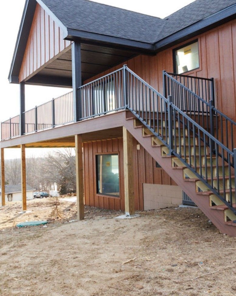 A wood-sided house with a raised deck, black metal railings, and an outdoor staircase leading down to a dirt yard.