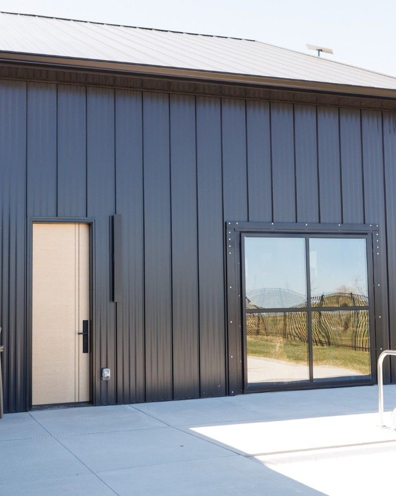 A modern building with dark vertical metal siding, a light-colored door, and a large black-framed square window.